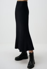 Jupe midi noire tricotée avec un ourlet évasé et une texture côtelée, associée à des bottes de combat noires à lacets. Silhouette élégante, sans motifs visibles.