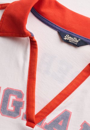 Camisa blanca con ribete naranja alrededor del cuello de pico y una etiqueta de la marca "SuperDry" en el interior del cuello.