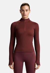 Top s dlhými rukávmi a zipsom na krku v hlbokom burgundskom odtieni, s vertikálnym rebrovaním a priliehavým strihom; obsahuje otvory na palce a kontrastné červené prúžky.