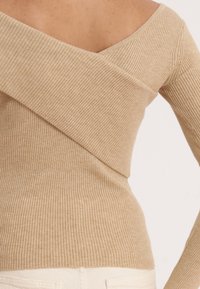 Beige värjätty neulepaita olkapäät paljastavalla mallilla ja vinoon kääritty yksityiskohta, pehmeällä tekstuurilla ja vartalonmyötäisellä siluetilla.