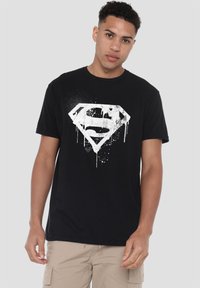 Black cotton t-shirt featuring a white splattered Superman logo design at the center, short sleeves, and a round neckline.