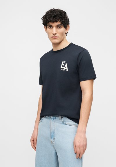 Νεαρός με σγουρά μαλλιά, που φοράει μαύρο μπλουζάκι με το λογότυπο EA και ανοιχτό μπλε τζιν, στέκεται μπροστά σε απλό φωτεινό φόντο.