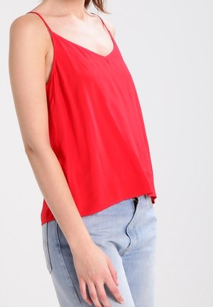 Kvinde iført en rød ærmeløs V-hals top kombineret med lyseblå denimjeans mod en ensfarvet baggrund.