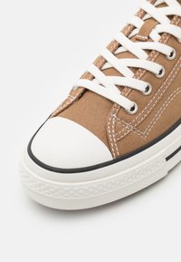 Sneaker in tela marrone con lacci bianchi, punta in gomma e rifiniture nere. Presenta cuciture a contrasto e una suola testurizzata per una migliore aderenza.