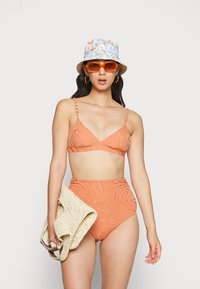 Biquíni laranja de cintura alta e textura, com padrão zigzag; top com alças ajustáveis. Modelo segura uma bolsa de palha beje e usa um chapéu de balde floral e óculos de sol laranja.