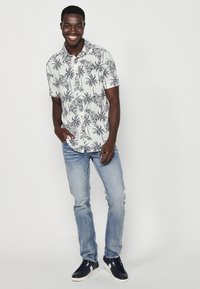 Maglietta a maniche corte con sfondo bianco e stampa di palme nere; abbinata a jeans azzurri e sneakers blu marine. Vestibilità dritta, design casual.