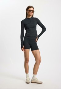 Top nero a maniche lunghe con collo alto, abbinato a pantaloni corti neri. L'outfit presenta una texture liscia e un design aderente, accessoriato con occhiali da sole.