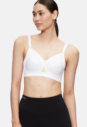 Femme aux cheveux foncés attachés en queue de cheval, portant un brassière de sport blanche et un legging noir taille haute, regardant vers sa gauche.