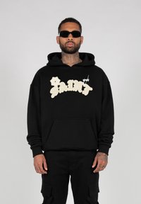 Hoodie noir avec "SAINT" en grandes lettres beiges, orné d'une accent floral. Le matériau semble doux ; comprend une poche avant et une capuche ajustable.