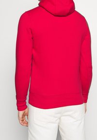 Felpa rossa in tessuto morbido e texturizzato, con tasca a marsupio e polsini a costine; indossata su pantaloni chiari.