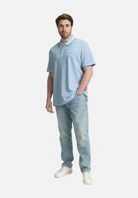 Ljusblå pikétröja med gröna detaljer och krage, kombinerad med ljusa denimjeans och vita sneakers, står med händerna i fickorna.