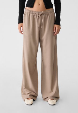 Vrouw draagt beige wijde broek met trekkoord in de taille, zwart croptopje en crèmekleurige platte schoenen, staand tegen een effen achtergrond.