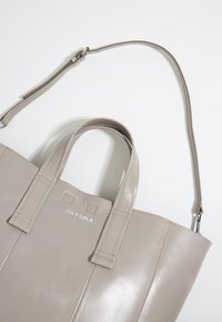 Borsa tote in pelle grigia con doppi manici e tracolla staccabile. Presenta una texture liscia e un logo impresso sulla parte anteriore.
