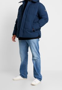 Marineblå polstret jakke med hætte, der har lynlåslukning og sidelommer. Kombineret med lyseblå jeans og hvide sneakers.