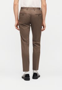 Ενήλικας που φορά στενά καφέ παντελόνια, λευκές κάλτσες και μαύρα επίσημα παπούτσια, εμφανιζόμενος από πίσω σε απλό λευκό φόντο.