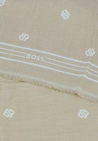 Sciarpa beige con una leggera texture, caratterizzata da motivi e marchi bianchi. Rifinita con bordi frangiati e dettagli a strisce bianche.