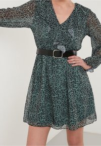 Vestido verde con estampado de leopardo, con escote en V fruncido, largas mangas transparentes, cintura ceñida con un cinturón negro y una falda fluida.