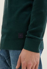 Suéter de punto verde texturizado con ribetes en el dobladillo y los puños, y un pequeño parche de logo negro en la parte inferior izquierda. Lleva puesto sobre unos jeans azules.