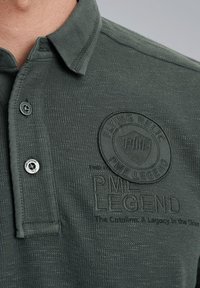 Donkergroene poloshirt van een gestructureerde stof, met een drieknoopsluiting, reliëflogo en een badge met de tekst "PME LEGEND".