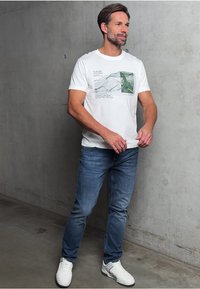 Vit grafisk t-shirt med grön bergdesign, tillsammans med blå jeans. Modellen har på sig vita sneakers och står mot en grå bakgrund.