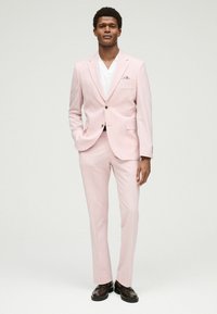 Helles rosa Anzug mit taillierter Passform, einreihiger Jacke, Reverskragen und slim-fit Hose. Kombiniert mit einem weißen Hemd und dunklen Slippern.