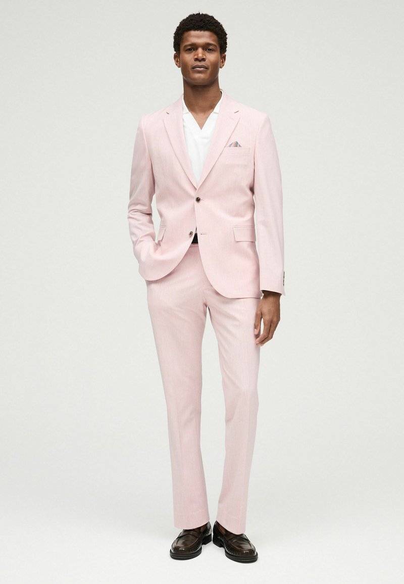 Helles rosa Anzug mit taillierter Passform, einreihiger Jacke, Reverskragen und slim-fit Hose. Kombiniert mit einem weißen Hemd und dunklen Slippern.