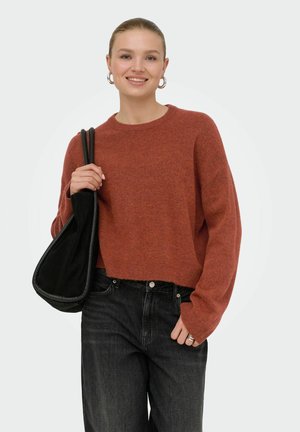 Jeune femme portant un pull couleur rouille et un jean noir, souriante, tenant un sac à épaule noir d'une main.