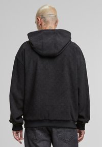Haut à capuche noir avec un motif en damier texturé, coupe oversize, capuche spacieuse et poignets côtelés. La vue arrière montre les détails des coutures et de la couture.