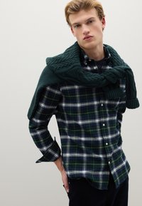 Camicia a quadri verde con accenti bianchi e gialli, design con bottoni frontali, abbinata a un maglione verde lavorato a maglia con trama, drappeggiato sulle spalle.