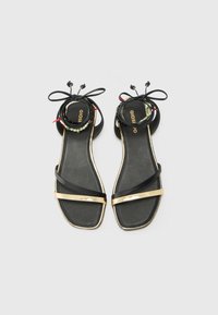 Sandalias planas negras con un diseño elegante, una tira metálica dorada y un lazo decorativo en el tobillo con cuentas de colores. Textura suave, forma cuadrada.