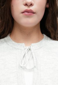 Lichtgrijze cardigan met een ronde hals, geribbelde textuur en een trekkoordsluiting aan de voorkant, gedragen over een witte top.