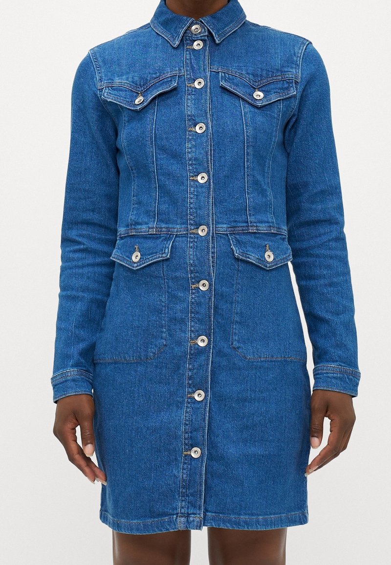 Femme portant une robe en jean bleue longueur genoux, à manches longues, avec des boutons sur le devant et des poches à rabat sur la poitrine et la taille.