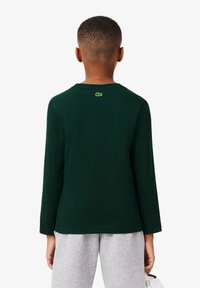 T-shirt verde a maniche lunghe in cotone, con scollatura rotonda e un piccolo logo ricamato sulla parte superiore della schiena. Indossato con pantaloni grigi.