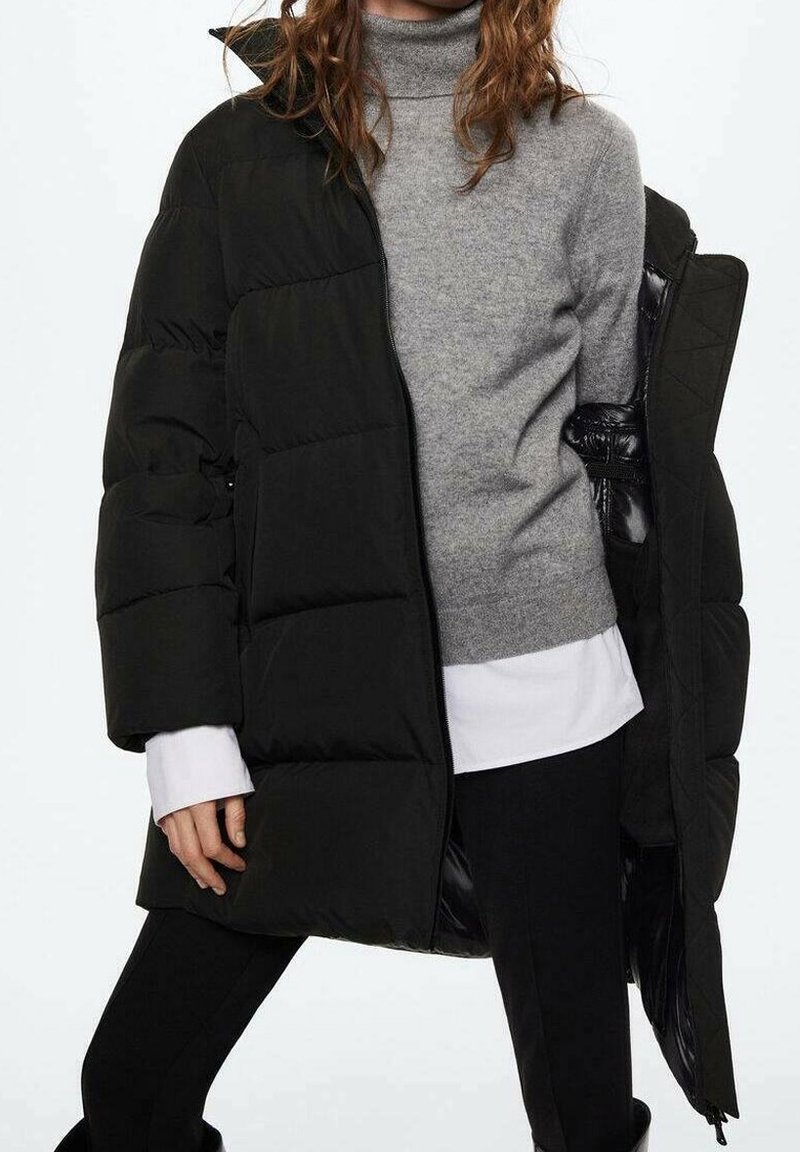 Femme portant un pull à col roulé gris sous un manteau matelassé noir, associé à un pantalon noir et une chemise blanche en-dessous.