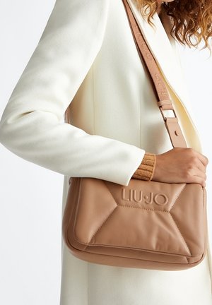 Sac bandoulière en cuir marron avec un design matelassé géométrique, logo embossé et sangle réglable. Porté sur un manteau de couleur crème.