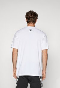 Balta medvilninė marškinė su trumpomis rankovėmis, apvalia iškirpte ir mažą juodą voro grafiką ant viršutinės nugaros dalies. Lygus paviršius, klasikinis įdaras.