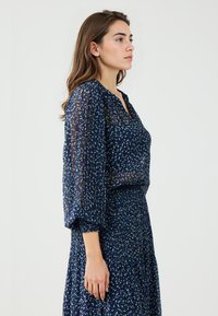 Navy blouse met een groen-witte stippenpatroon, voorzien van een V-hals en lange, geplooide mouwen; gelaagd met een bijpassende geplooide rok.