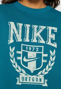 Turkos Nike sweatshirt med stora slitna vita grafiker, en skölddesign, lagerblad och texten "1972 Oregon". Rundad halslinje.