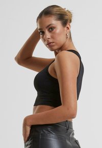 Crni uski top s tank žicom, napravljen od glatkog materijala. Model pozira s jednom rukom na svom boku, noseći crne kožne hlače.