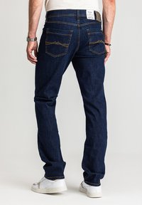 Ciemnoniebieskie dżinsy z prostą nogawką, posiadające dwie tylne kieszenie z pomarańczowym przeszyciem oraz brązową skórzaną metkę na pasku.