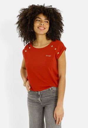 Mujer joven con cabello rizado que lleva una blusa roja de manga corta con botones dorados y el texto "Sé feliz", combinada con jeans grises y joyería dorada.