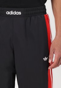 Pantaloni sportivi neri con strisce laterali rosse, vita elastica e logo adidas ricamato bianco sulla coscia sinistra. Texture liscia.
