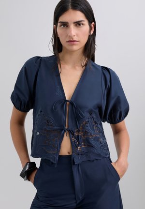 Blusa corta azul oscuro con mangas abullonadas, que presenta recortes de encaje y diseños florales bordados, atada en la parte delantera con una cinta.