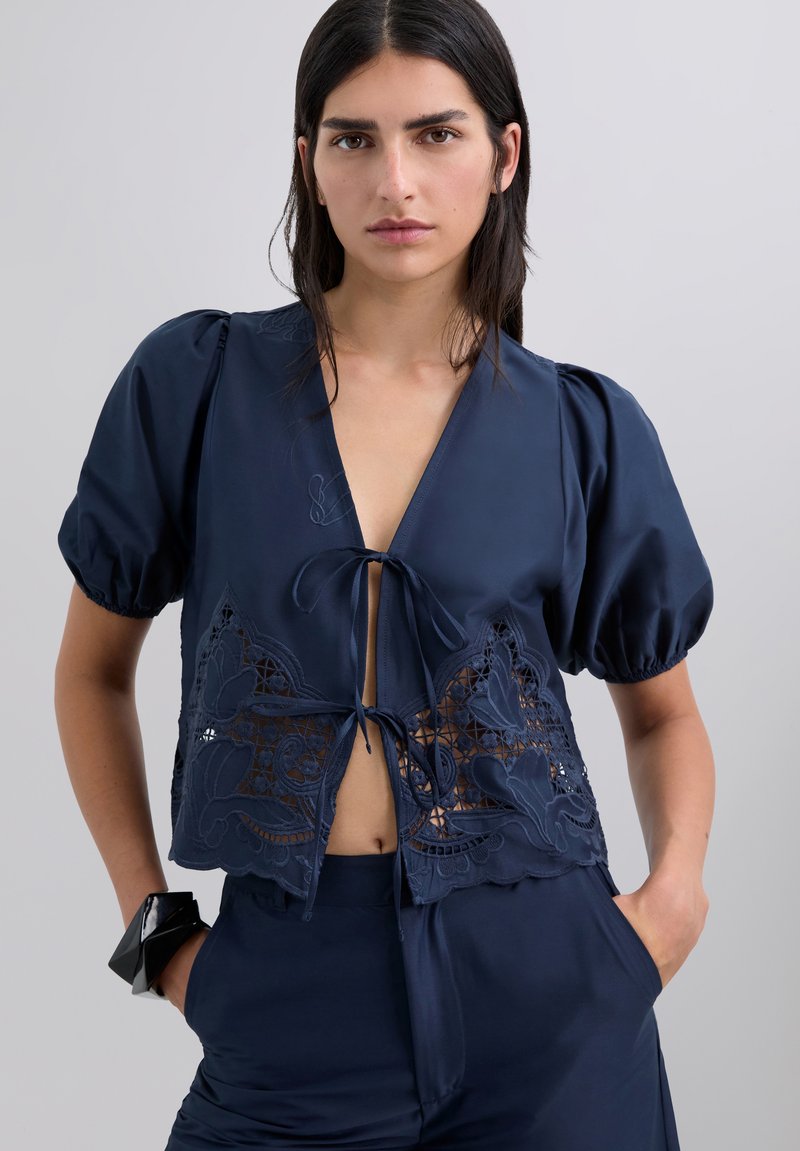 Blusa corta azul oscuro con mangas abullonadas, que presenta recortes de encaje y diseños florales bordados, atada en la parte delantera con una cinta.