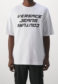 Bílý bavlněný tričko s odvážným černým grafickým potiskem, na kterém je diagonálně napsáno "VERSACE JEANS COUTURE".
