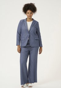 Completo blu su misura con blazer a singolo bottone e pantaloni a gamba larga. Il tessuto presenta una sottile trama, abbinato a una maglietta leggera e scarpe bianche.