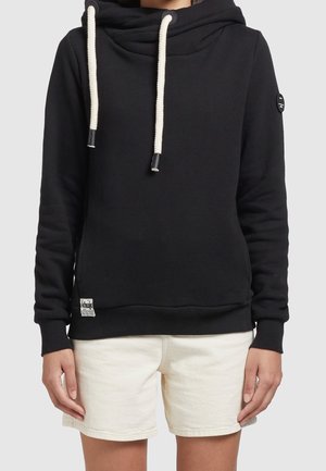 Osoba ubrana w czarną bluzę z kapturem z grubymi białymi troczkami i kremowe szorty, stojąca na tle jednolitego jasnoszarego tła.