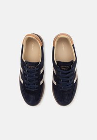 Navy blue suede sneakers with white stripes, tan accents, round toe, and navy laces. Gold logo on the insole. Smooth textured sole.