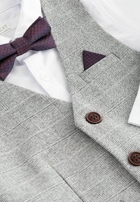 Szürke texturált öltönymellény, két barna gombbal, fehér inggel és sötétvörös mintás született csokornyakkendővel párosítva. Zsebkendő is tartozik hozzá.