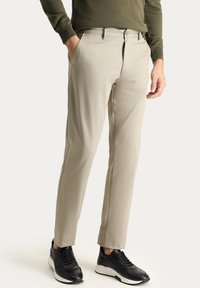 Pantalones de mezcla de algodón color beige con un corte ajustado, que cuentan con una textura suave, pasadores para cinturón y bolsillos frontales, combinados con zapatillas negras.